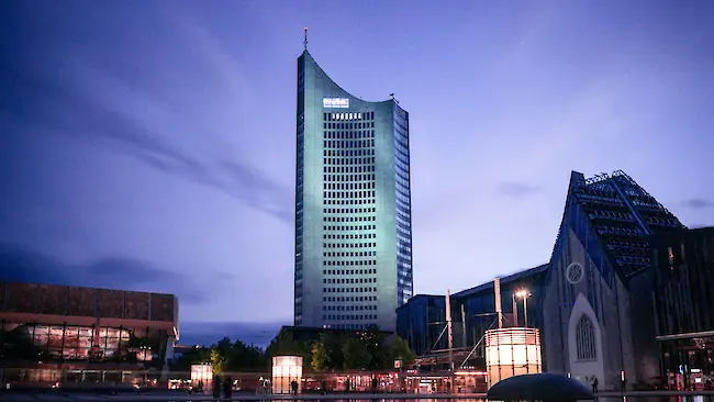 Blue-hour evening view in Leipzig with reflections of a lit high-rise and the university building on a central pond. Blue-hour evening view in Leipzig with reflections of a lit high-rise and the university building on a central pond.